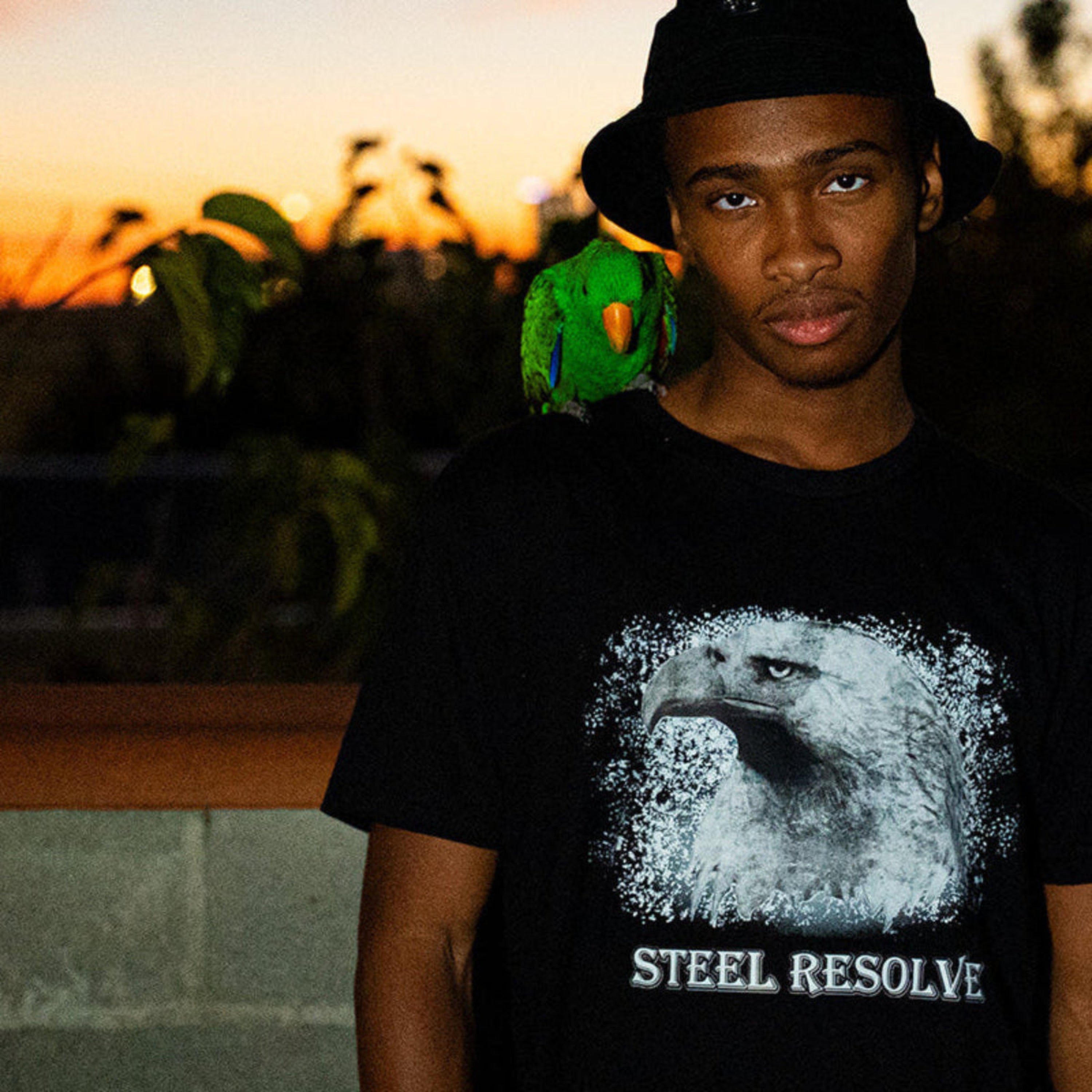 Wisdom wearing a black t-shirt with an eagle design and 'Steel Resolve' text, standing outdoors at sunset.